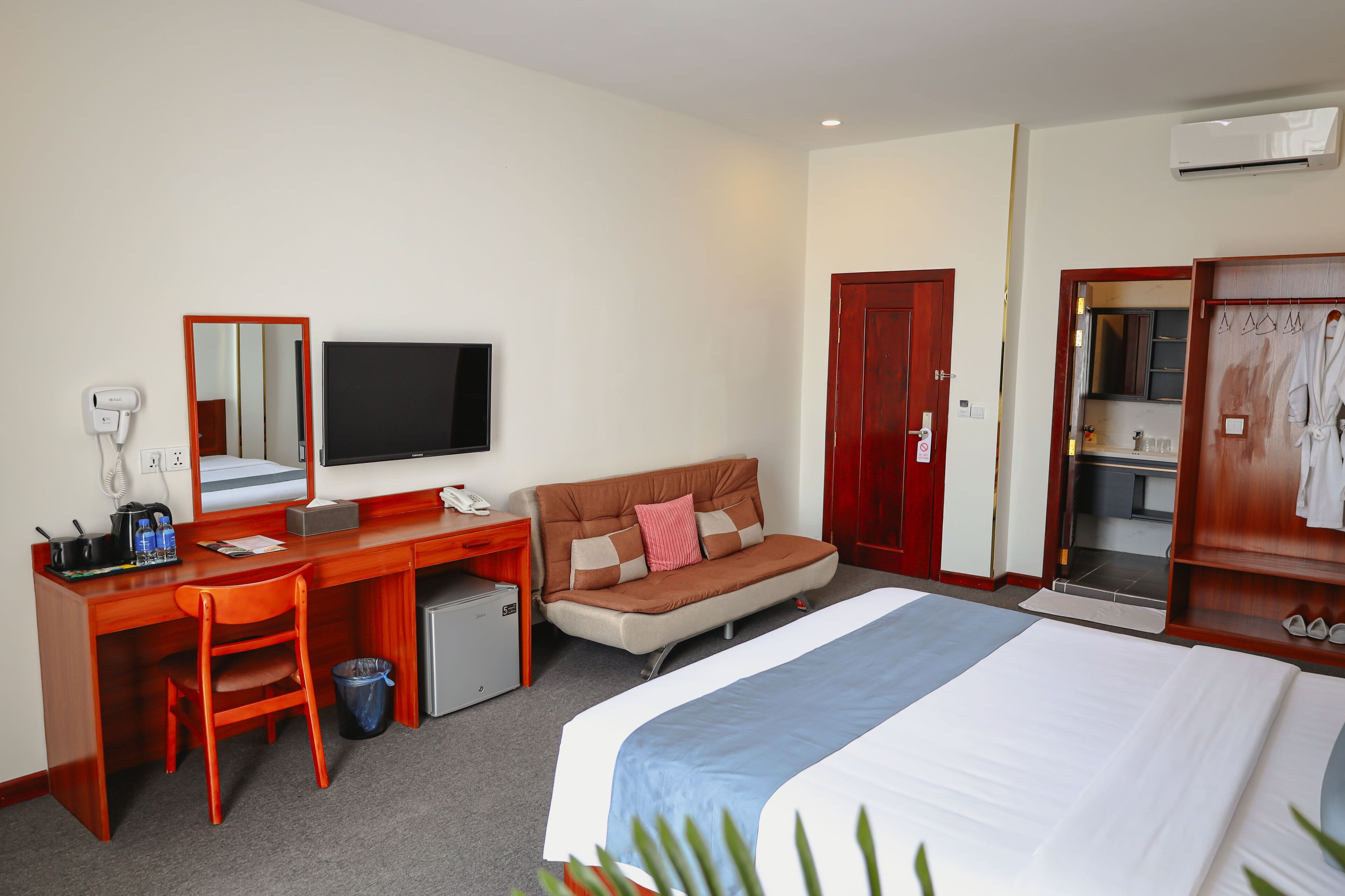 A hotel room with a bed, a desk with a computer, a chair, a small fridge, a TV, and a wardrobe.