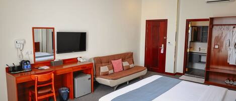A hotel room with a bed, a desk with a computer, a chair, a small fridge, a TV, and a wardrobe.