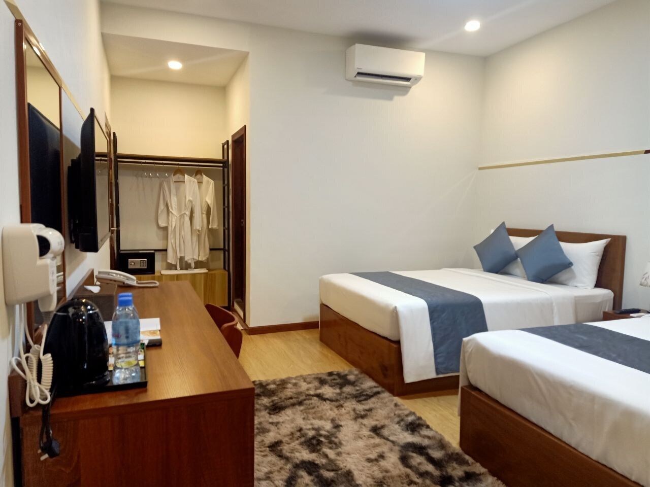 A hotel room with two single beds, a wooden desk with a telephone and a water bottle, a mirror, and an air conditioning unit.