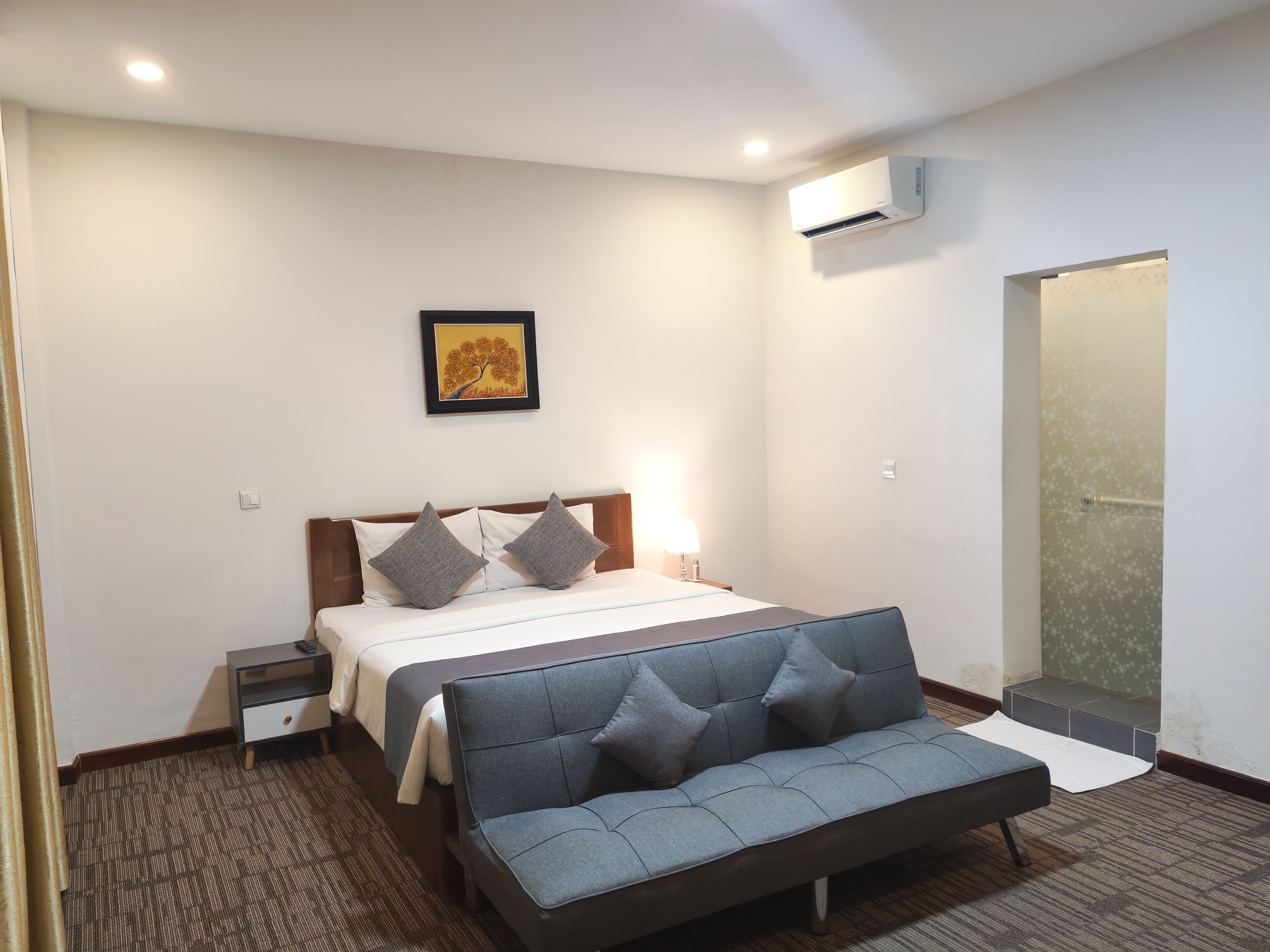 Habitación de hotel con cama, mesitas de noche, sofá, equipo de aire acondicionado y un cuadro en la pared.