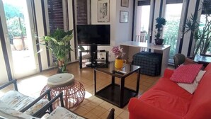 Un saló amb un sofà vermell, una televisió i un ventilador de sostre. Hi ha plantes i una finestra amb persianes.