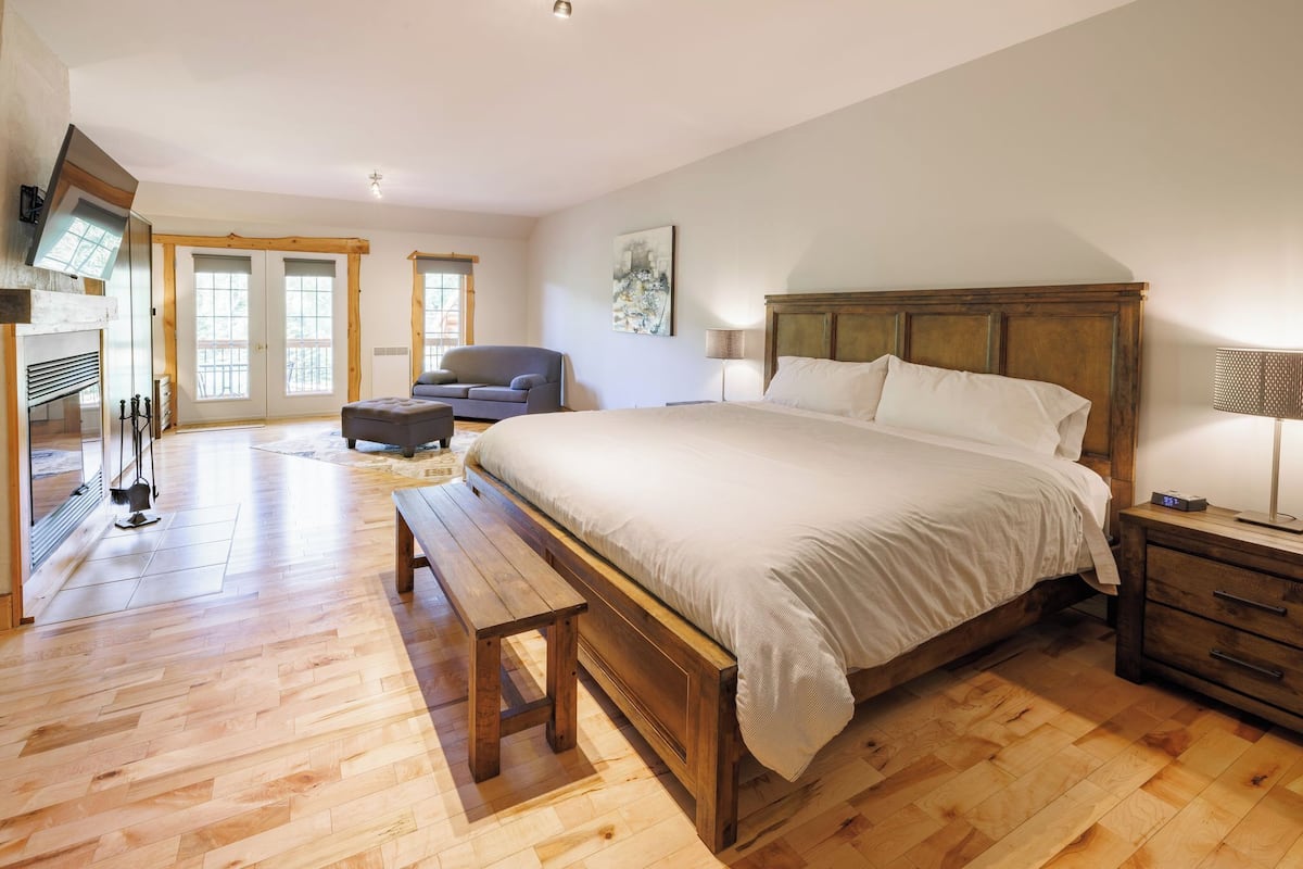 Um quarto espaçoso com uma cama grande, piso de madeira, um banco, uma cadeira, uma televisão e uma lareira.