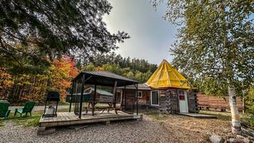 A rustic cabin with a wooden deck and a yellow awning, surrounded by trees and gravel.