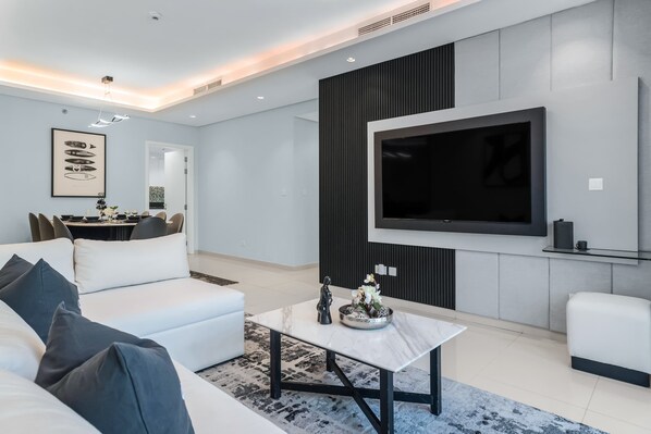A modern living room with a white sofa, a coffee table, a flat-screen TV mounted on the wall, and a dining area in the background.