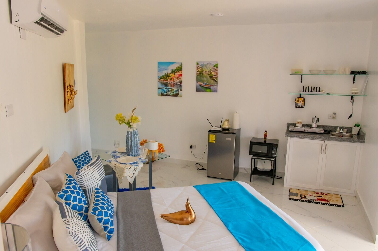 A compact hotel room with a bed, a small dining table, a kitchenette, and a single artwork on the wall.