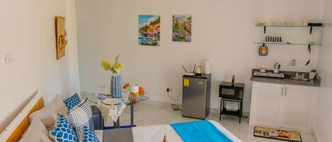 A compact hotel room with a bed, a small dining table, a kitchenette, and a single artwork on the wall.