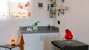 Cocina compacta con refrigerador, pileta y una pequeña mesada. Se observa una ventana con cortina estampada de frutas, un estante con utensilios de cocina y un objeto decorativo rojo sobre la mesada.