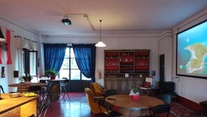 A room with a wooden table, chairs, a bar area with shelves, a projector screen, and a large window with curtains. - The Lime Tree (Castiglion Ubertini)