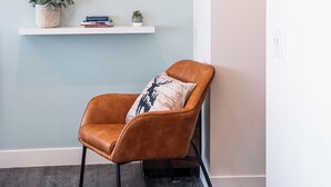 Una habitación moderna con un sillón de cuero marrón, dos estanterías blancas con plantas y libros, y una pared con cuadros enmarcados.