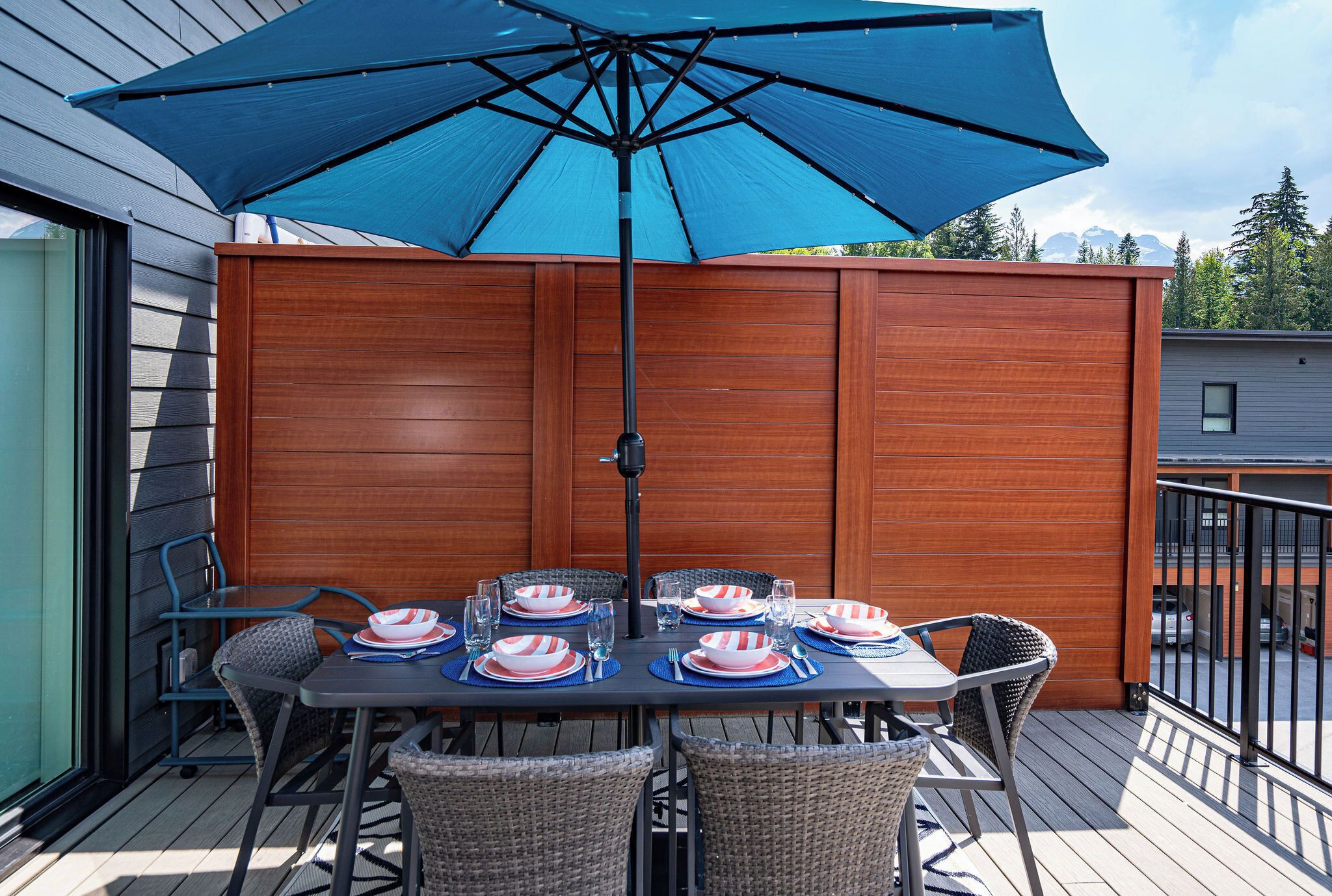An outdoor dining area with a table set for four, a blue umbrella, and a wooden privacy screen.