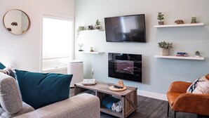 Una sala de estar moderna con un televisor de pantalla plana, una mesa de centro, un sofá y una silla. Se observan estanterías con plantas y objetos decorativos en la pared.