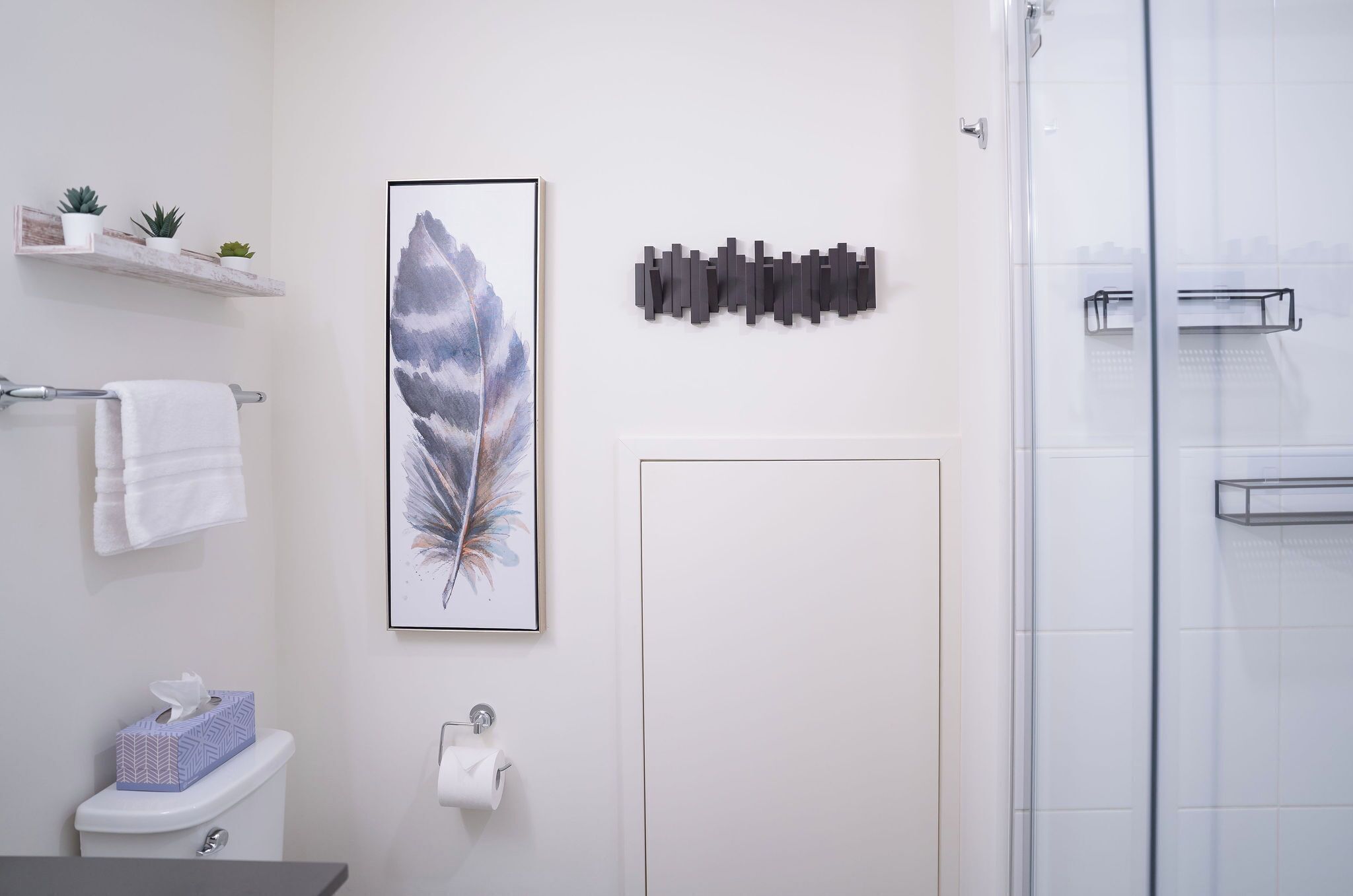 A bathroom with a toilet, a towel rack, a framed feather artwork, a small shelf with plants, and a shower area.