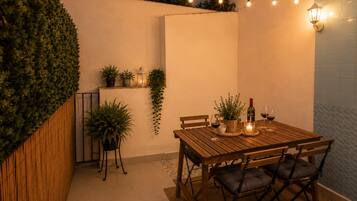 Una mesa de madera con sillas, una vela y una botella de vino, rodeada de plantas en macetas y luces de cuerda.