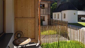 Uma pérgola de madeira com uma mesa e cadeiras, um edifício branco com telhado vermelho e um jardim cercado.