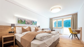 Habitación de hotel moderna con una cama grande, cabecera de madera, una mesita de noche, un escritorio y una silla. Se observa un ventanal con cortinas, un cuadro en la pared y una lámpara del techo.
