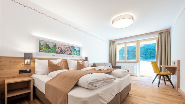 Quarto de hotel moderno com uma cama grande, cabeceira de madeira, uma mesa de cabeceira, uma escrivaninha e uma cadeira. Há uma janela grande com cortinas, um quadro na parede e uma luminária no teto.