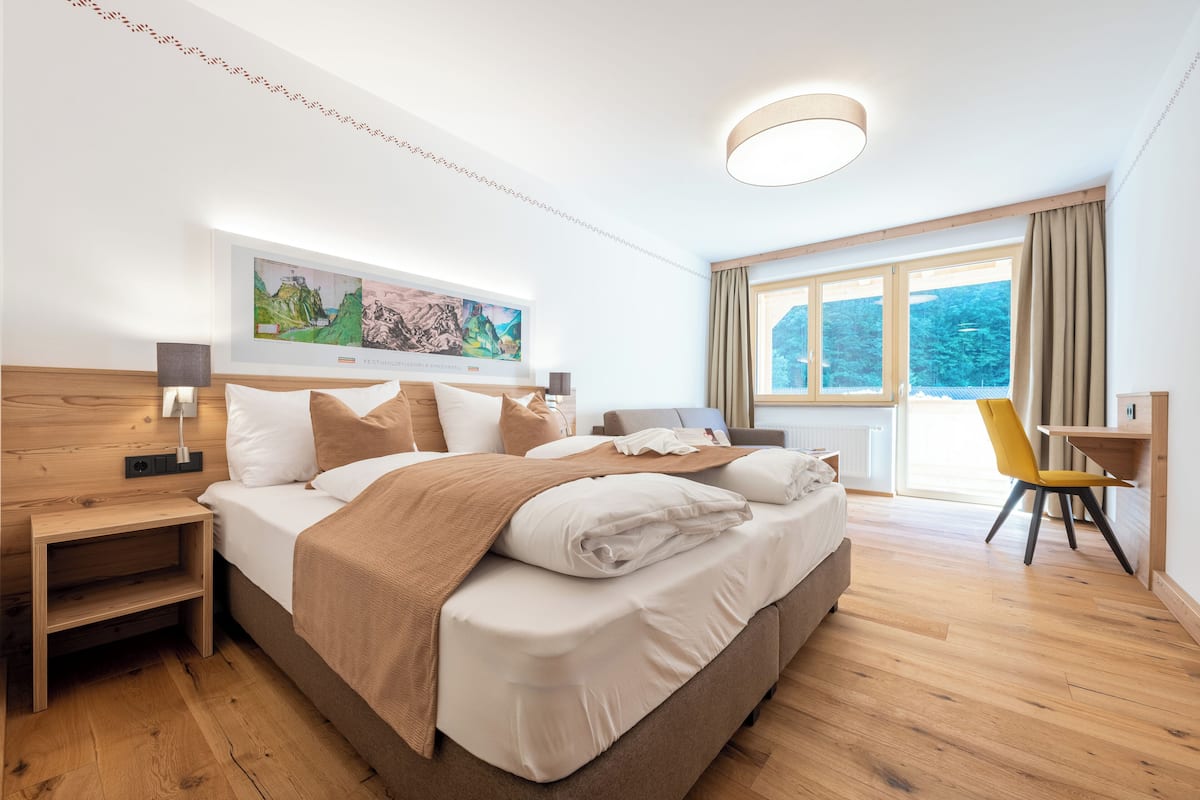 Habitación de hotel moderna con una cama grande, cabecera de madera, una mesita de noche, un escritorio y una silla. Se observa un ventanal con cortinas, un cuadro en la pared y una lámpara del techo.