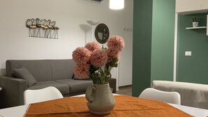 Een moderne woonkamer met een grijze bank, een witte salontafel met een bruine tafelkleed, en een vaas met roze bloemen.