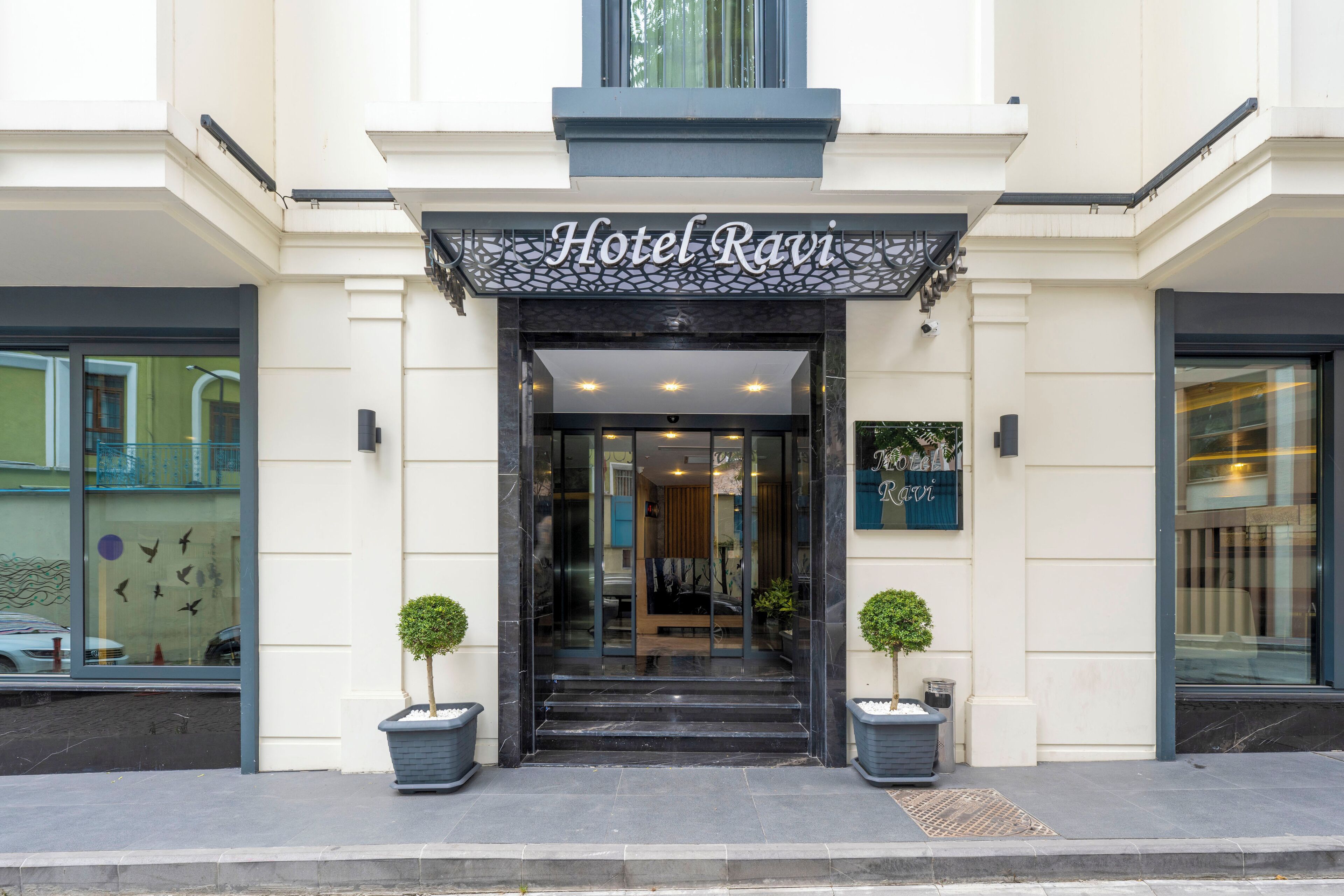 La entrada del Hotel Ravi, con una fachada moderna, puertas de vidrio y un cartel sobre la entrada.