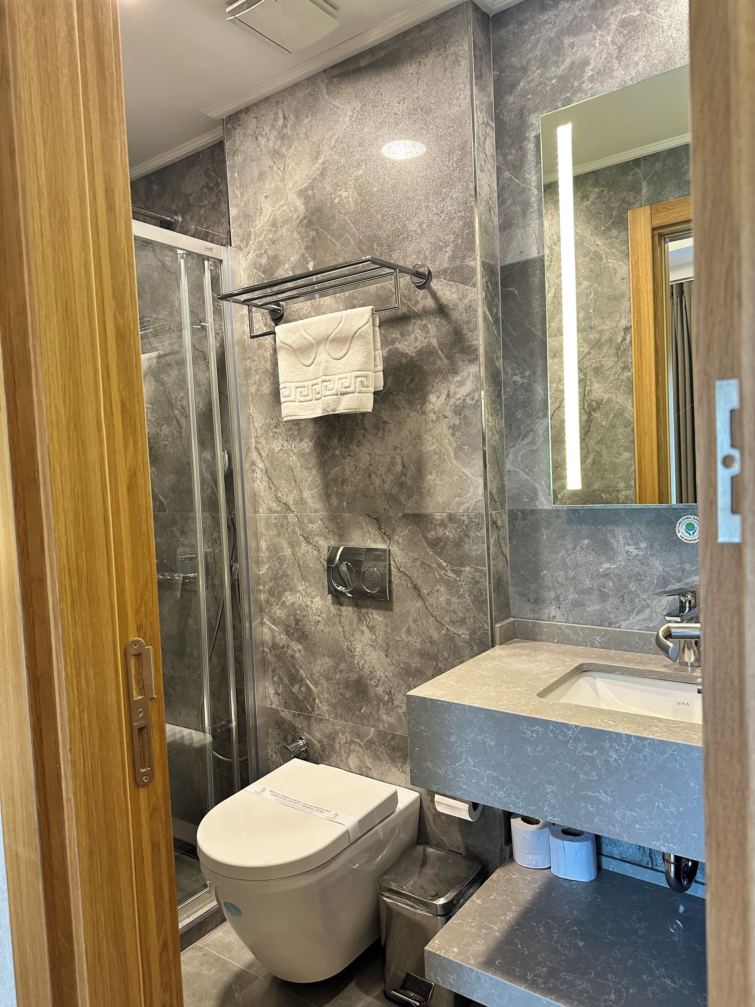 A modern bathroom with a marble wall, a glass shower enclosure, a toilet, and a sink with a mirror above it.
