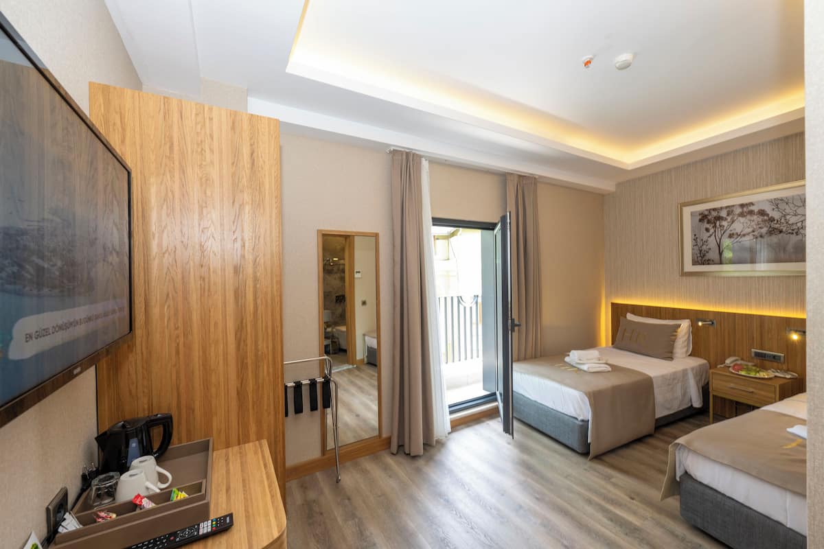 Habitación de hotel con piso de madera, dos camas, un escritorio con cafetera, televisor y puerta corrediza a un balcón.