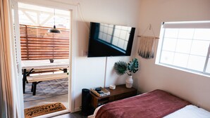 Ein Schlafzimmer mit einem Bett, einem an der Wand befestigten Fernseher, einem Nachttisch mit Pflanze und einem Fenster mit Jalousien.