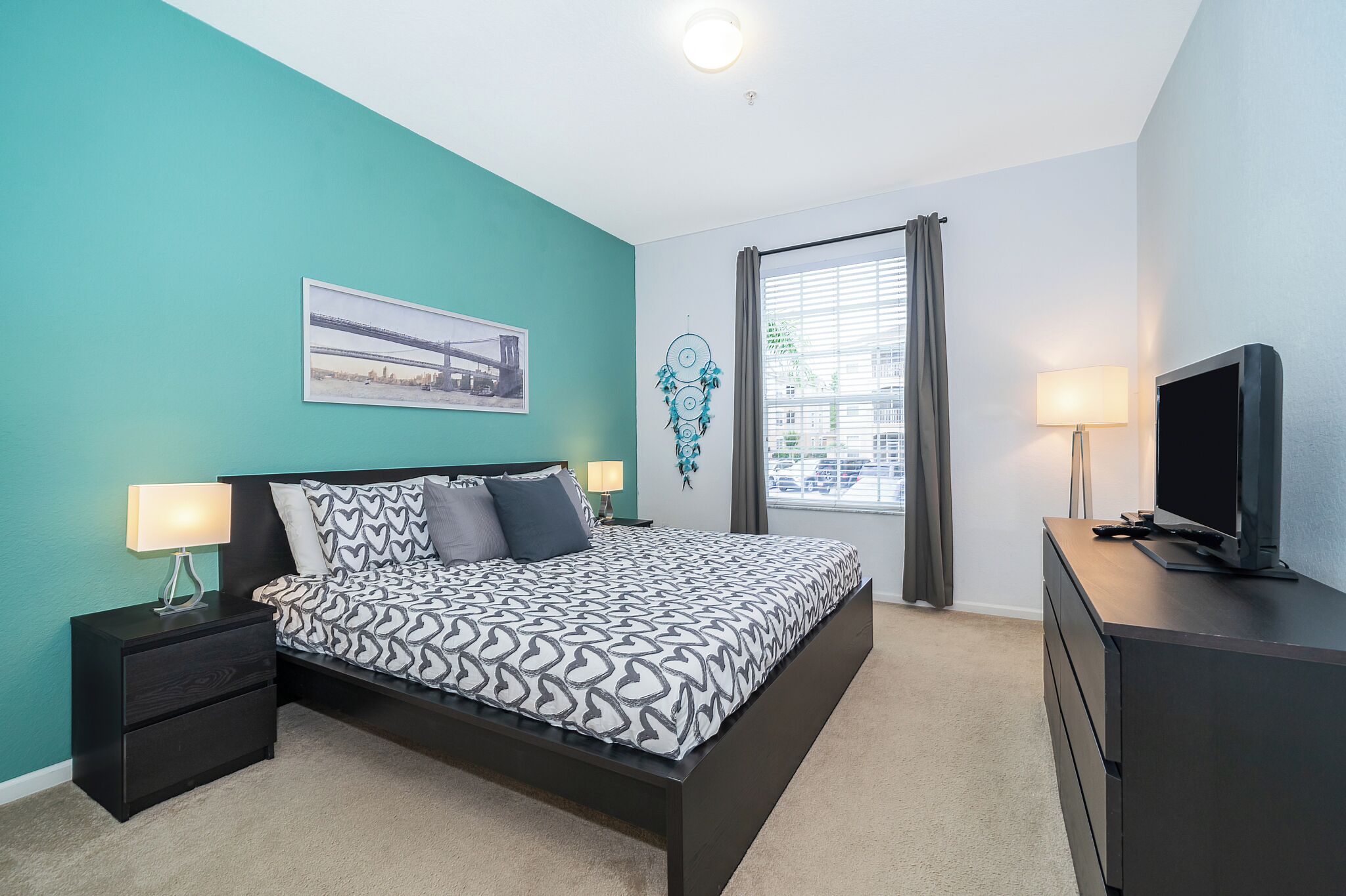A modern bedroom with teal walls, a large bed with patterned bedding, two bedside tables with lamps, a TV on a dresser, and a window with curtains.