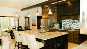 A modern kitchen with dark cabinetry, a large island with bar stools, pendant lighting, and a view of the dining area.