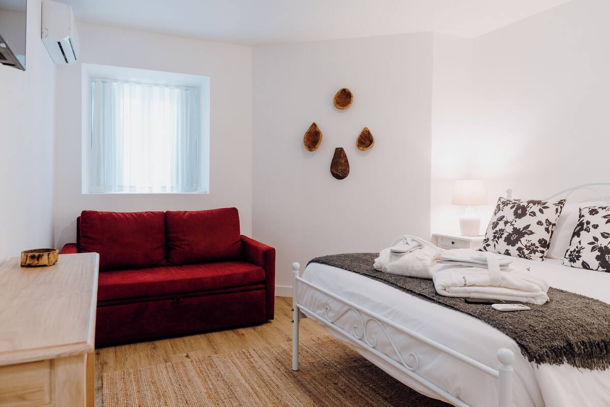 A bedroom with a bed, a red sofa, a window, and wall art.