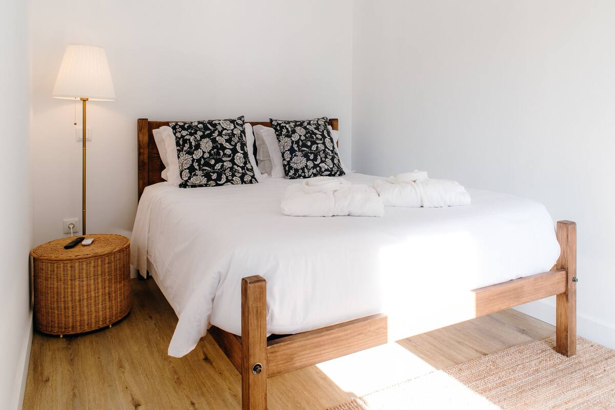 A neatly made bed with white linens, two floral-patterned pillows, and a remote control on a wicker side table.
