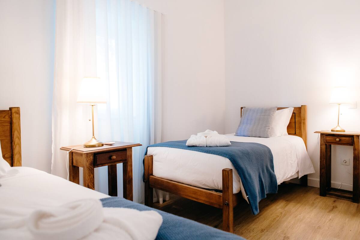 A hotel room with a wooden bed, a bedside table with a lamp, and a window with sheer curtains.