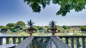 Une balustrade en pierre surplombe une rivière bordée d’arbres, avec un bâtiment visible au loin.
