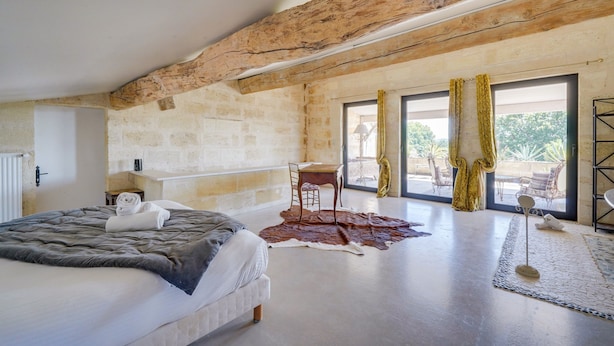 Une chambre avec un grand lit, un plafond en bois, une petite table avec une chaise, et une vue sur l’extérieur grâce à de grandes fenêtres.