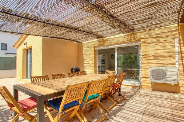 Um pátio externo com mesa e cadeiras de madeira, churrasqueira e porta de correr que dá para um espaço interno.