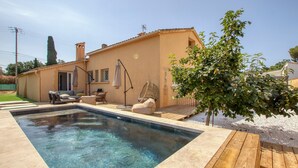 Une piscine avec une terrasse en bois, un espace de détente comprenant une chaise et un parasol, et un bâtiment doté d’un toit en tuiles.