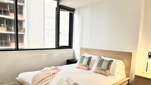 Una cama bien hecha con almohadas, una colcha y toallas enrolladas. Hay una mesita de noche con una lámpara y un velador.