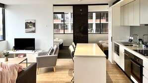 Una cocina moderna con armarios blancos, una isla central y un horno empotrado. Hay un área de comedor con un sofá y un televisor.