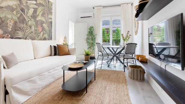 Ein modernes Wohnzimmer mit einem weißen Sofa, einem Couchtisch und einem Fernseher. Es gibt einen Essbereich mit Stühlen und einem Tisch, sowie ein großes Fenster mit Vorhängen.