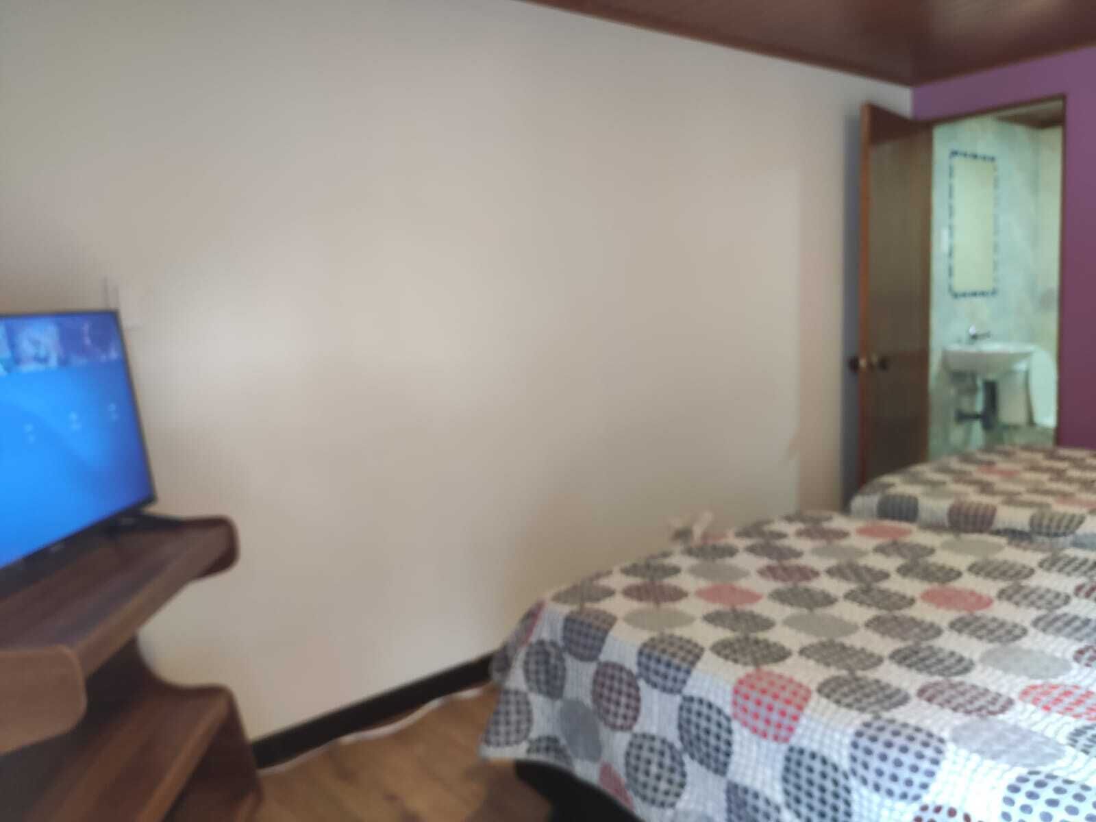 A bedroom with a bed, a television on a wooden stand, and a bathroom visible through an open door.