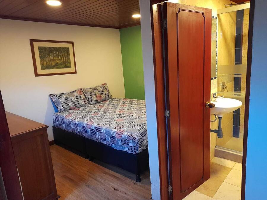 Un dormitorio con cama, armario de madera, pared verde y baño con lavamanos y espejo.