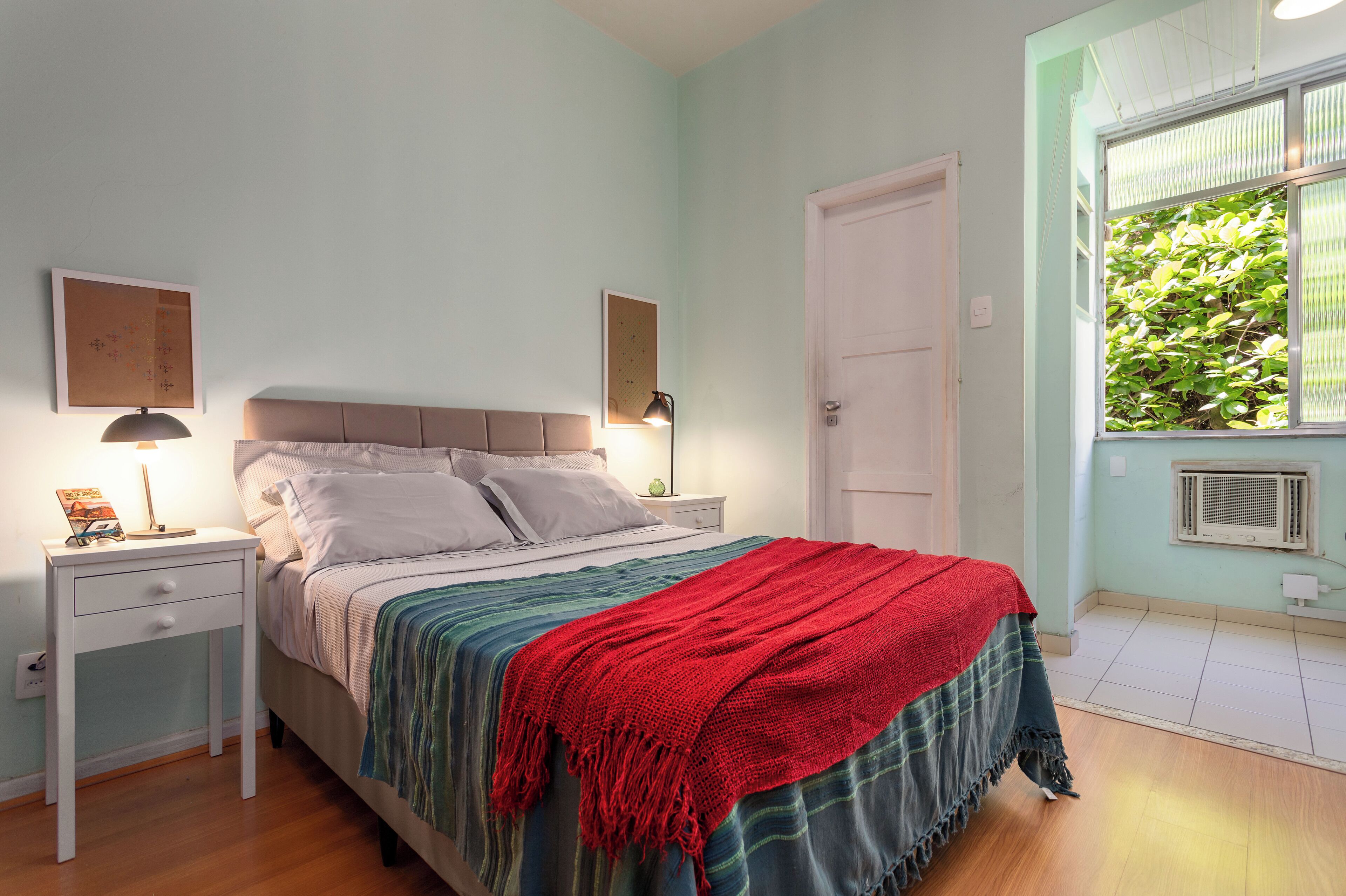 A bedroom with a bed, bedside tables, a window with a view of greenery, and a door.