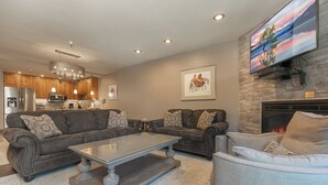A modern living room with a fireplace, a large flat-screen TV mounted above it, and a kitchen in the background.