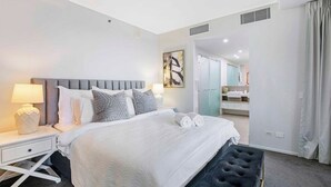 Une chambre d’hôtel moderne avec un grand lit, une table de chevet avec une lampe, un pouf noir et une salle de bain visible à travers une porte ouverte.