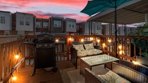 Een terras met rieten meubels, een barbecue en sfeerverlichting tegen een zonsondergang.