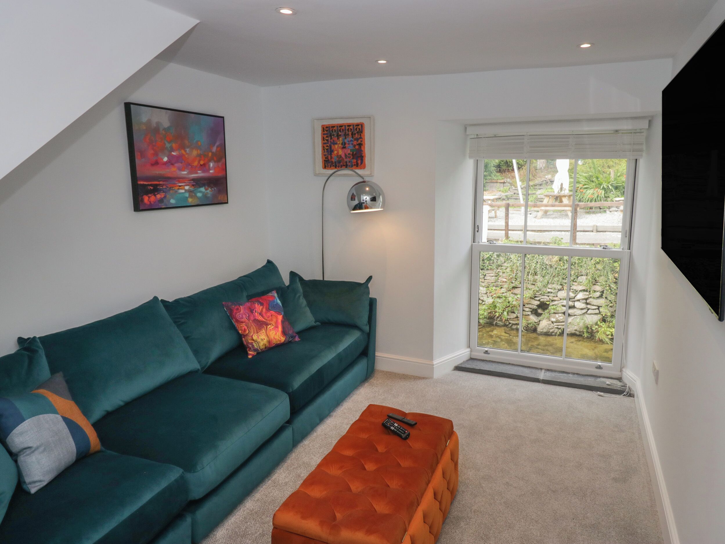 Una sala de estar moderna con un sofá azul verdoso, un pequeño taburete naranja y un ventanal con vistas a una pared de piedra y vegetación.