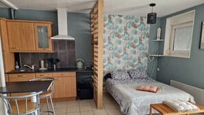 A compact studio apartment with a kitchenette, a bed with a patterned headboard, a small wooden table, and a window with blinds.