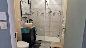 A bathroom with a toilet, a sink with a mirror, a shower, and a small table.
