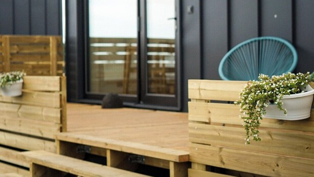 Un edificio moderno con fachada oscura, puerta corredera de vidrio y bancos de madera con plantas en macetas.