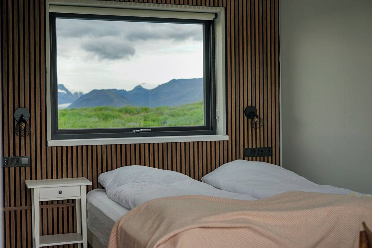 A bedroom with a large window, a bed, a bedside table, and a wall-mounted lamp.