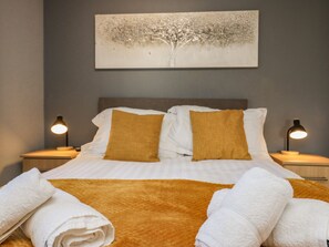 Een netjes opgemaakt bed met witte en mosterdkleurige kussens, een bruin plaid, en twee witte handdoeken. Aan de muur hangt een schilderij van een boom, en aan weerszijden van het bed staan twee nachtlampen.